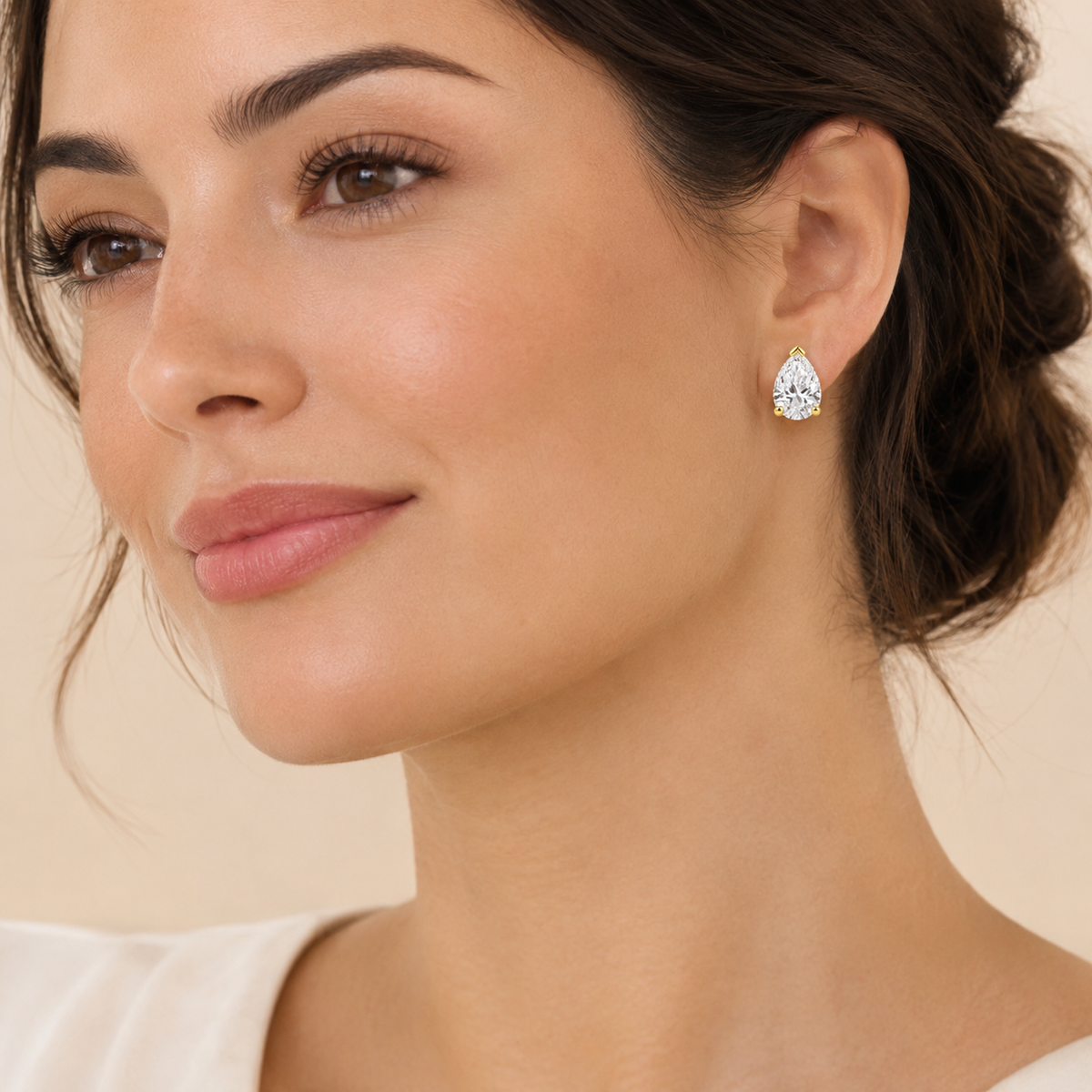 A close-up of a woman wearing a single pear-cut diamond stud earring with a silver-toned setting against a soft beige background.